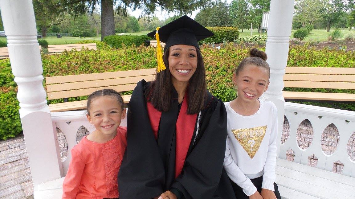 Shayla and daughters at graduation ceremony. Shayla in black robe with red sash. Girls seated in white gazebo at park.